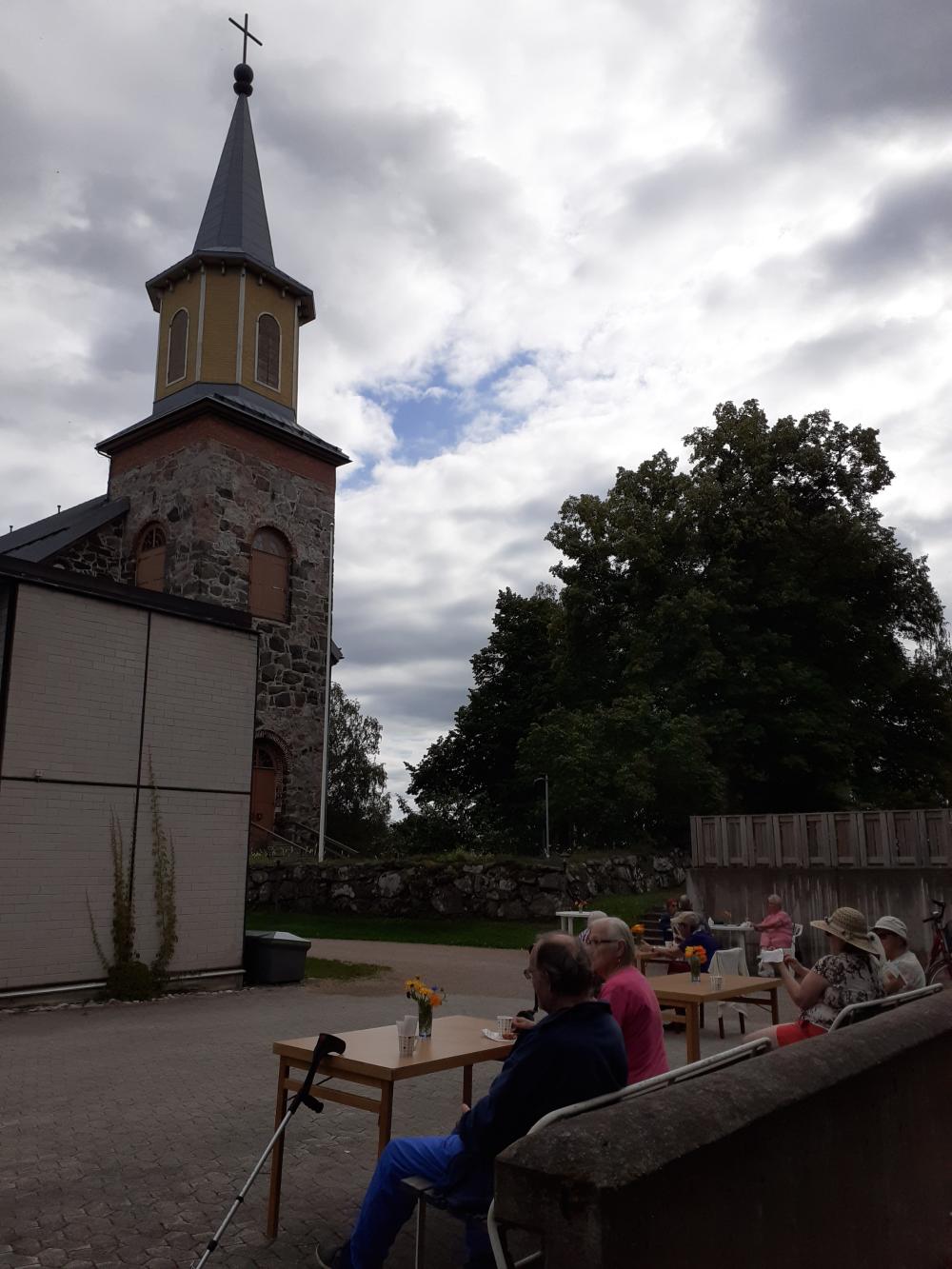 Kuvassa taustalla Karjalohjan kirkko ja kellotorni, pilvinen kesätaivasja ihmisiä ulkona kahvilla.