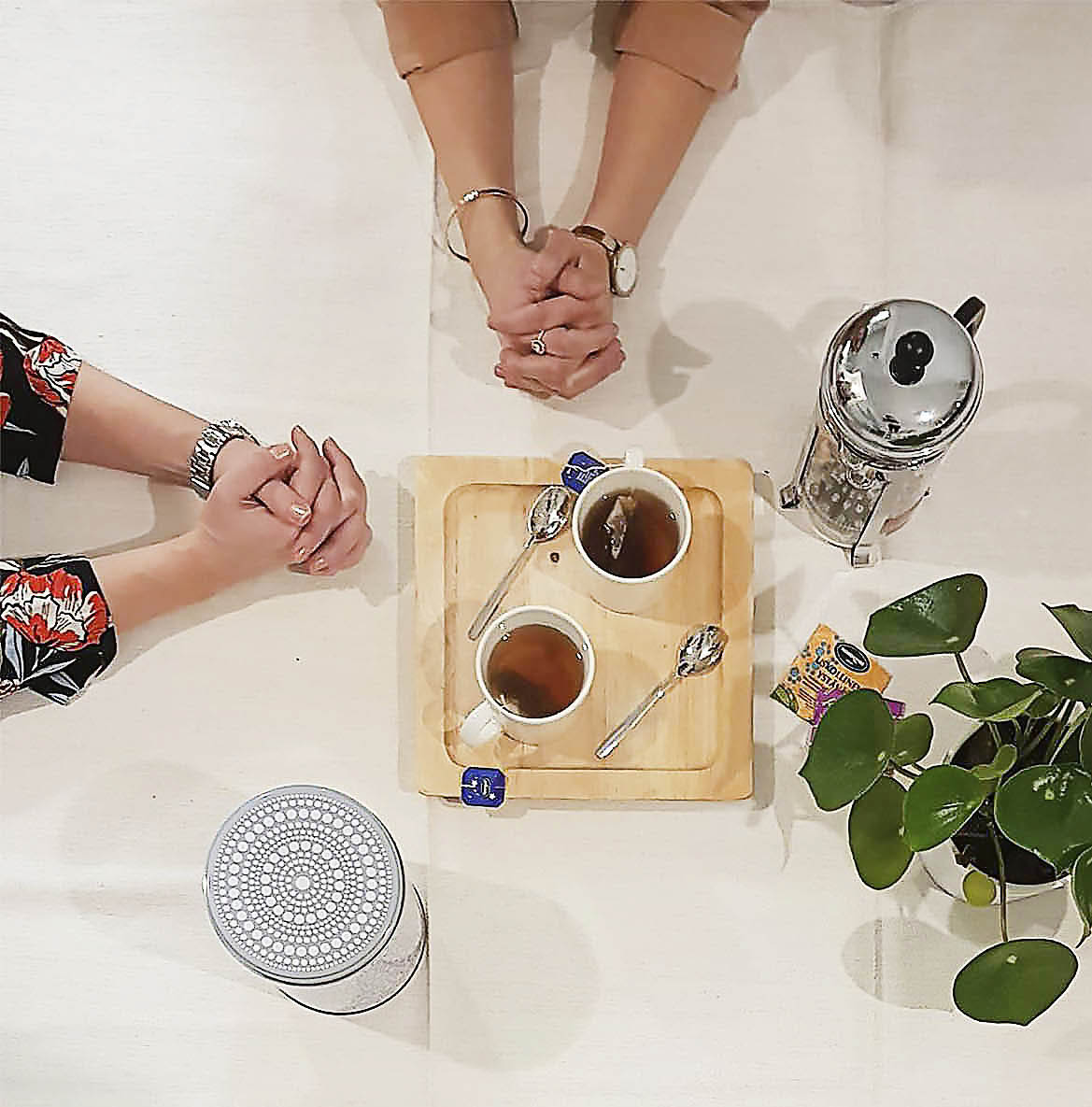 kahden naisen kädet ristissä pöydän päällä teekupit ja kannu sekä viherkasvi