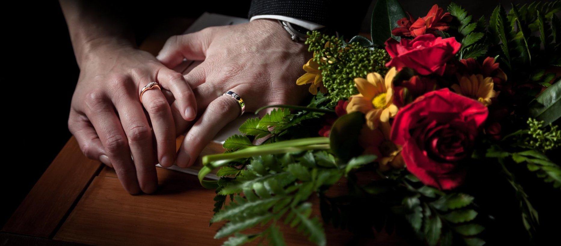 Hääpari pitää toisiaan kädestä kiinni sormukset sormissaan. Etualalla myös kukkia.