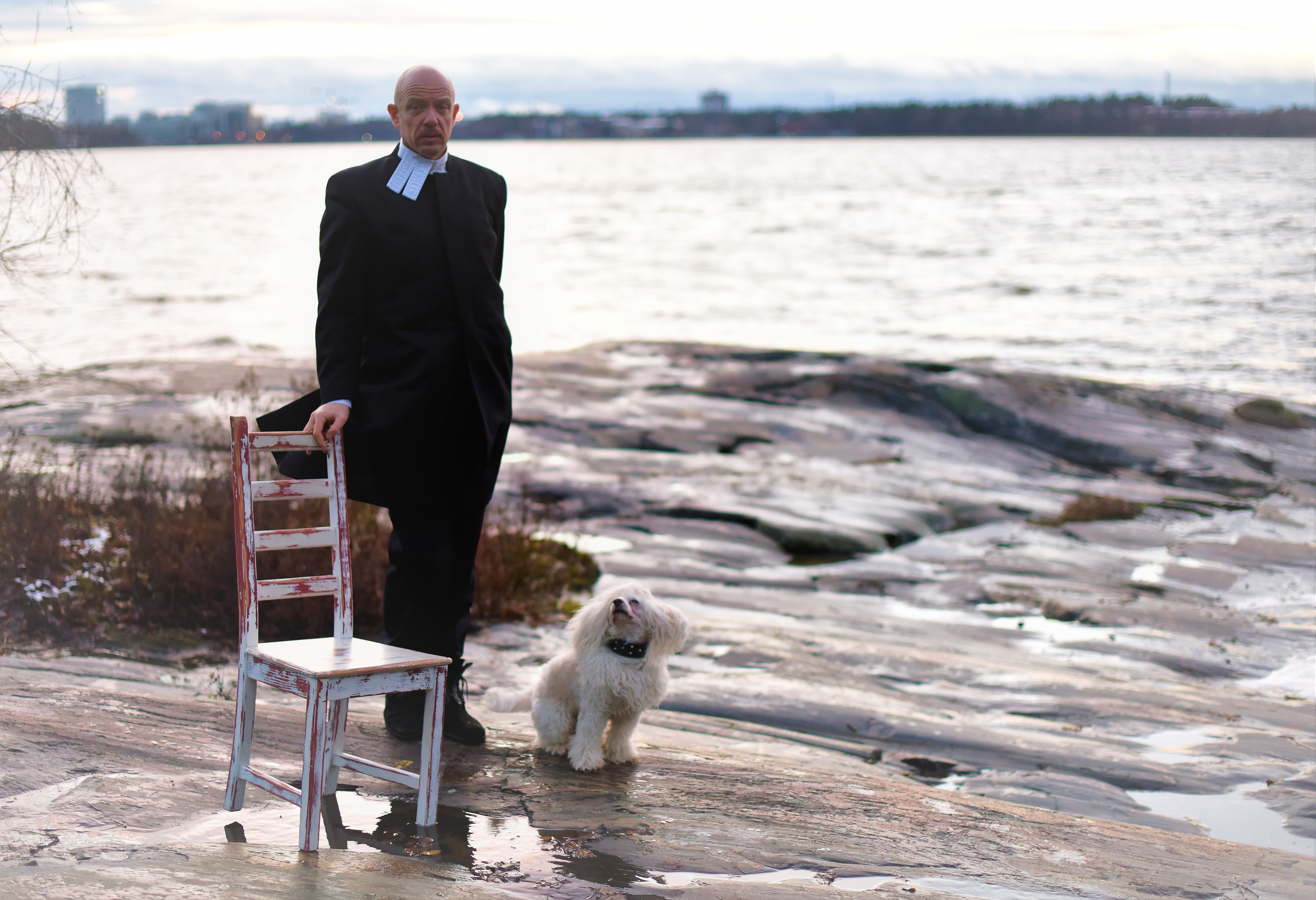 Lassi Alhorinne seisoo meren rannalla papinkaapu yllään vierellään pieni valkoinen Sielu-koira ja tuoli