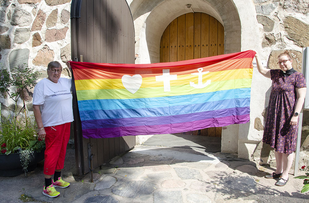 Naispastorit pitelevät sateekaarilippua Pyhän Laurin kirkon ovella kesäisissä maisemissa.