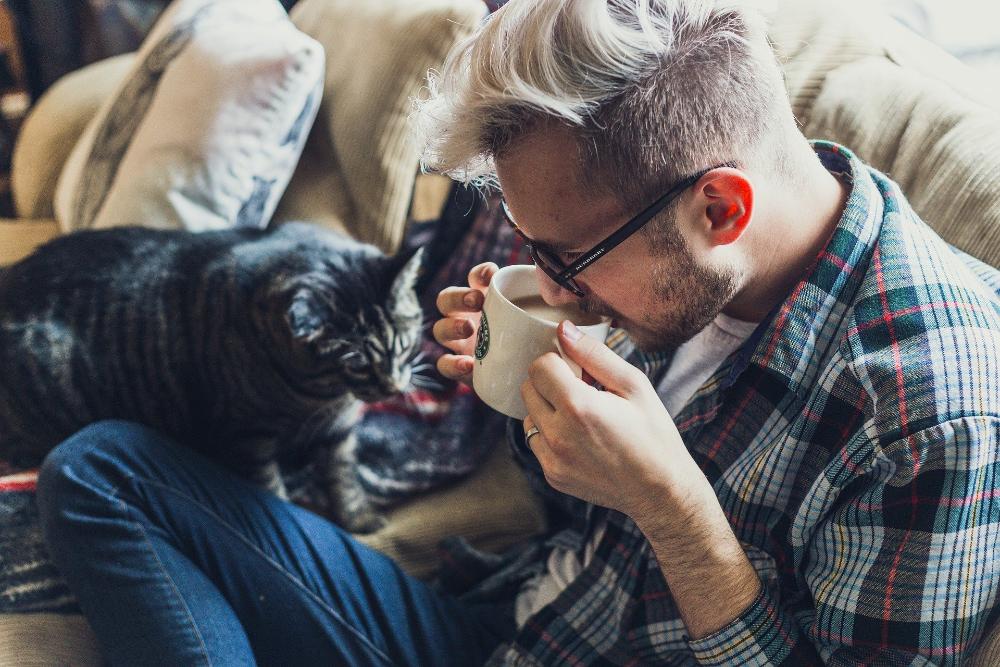 Mies juo kahvia sohvalla samalla kun kissa katselee vieressä